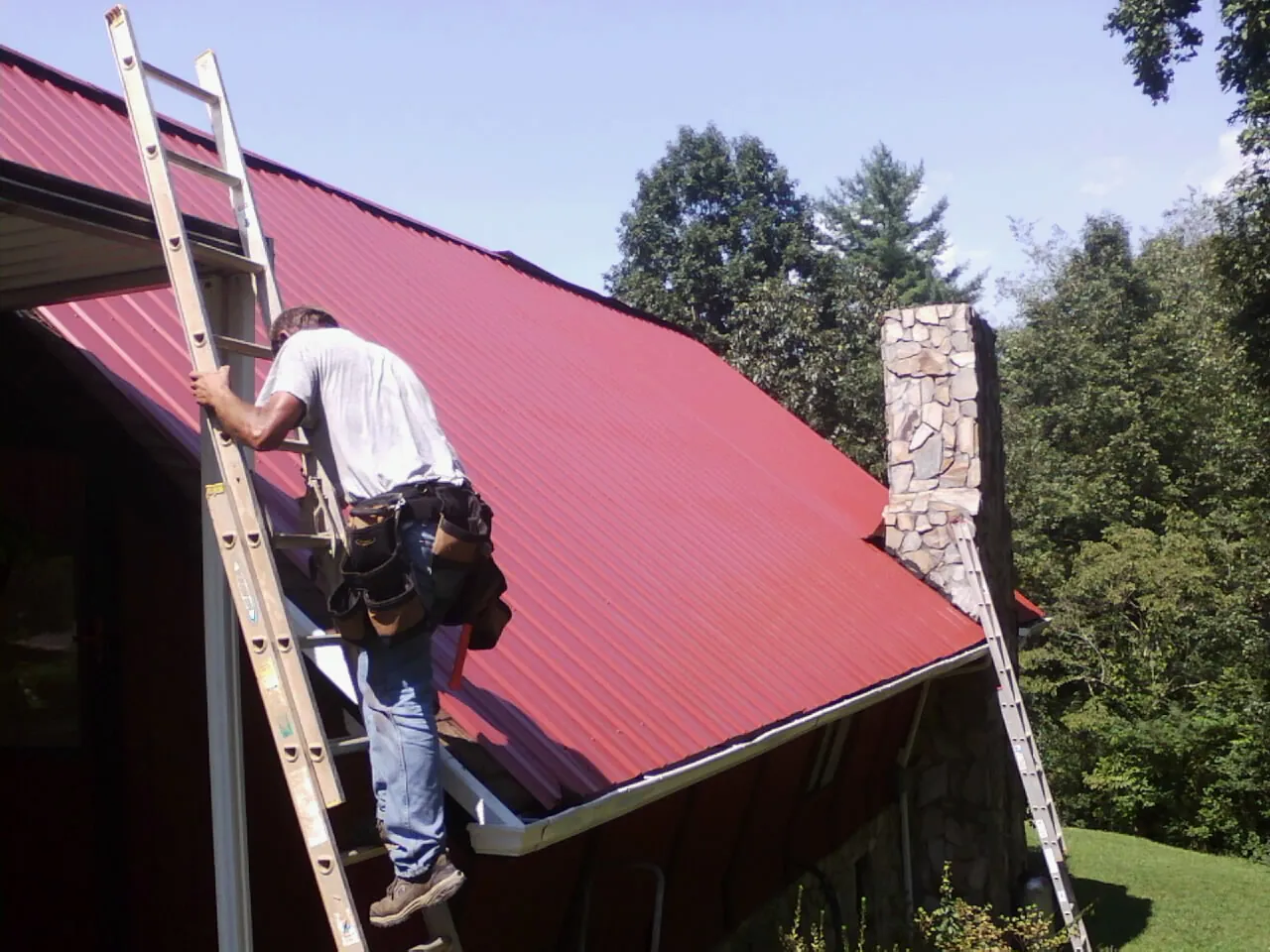 Roofer on a ladder beside a red metal roof and stone chimney — Soffit & Fascia Repair by Roofing Brothers in Braselton